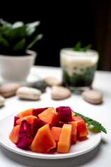 Slices of papaya and dragon fruit on a white plate with ice cendol background photographed on a white table