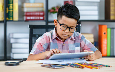 Smart Asian boy wearing glasses and pink checked shirt sitting on desk of magnifying glass, smartphone, and colorful pencils in reading room and concentrate on tablet study to complete homework