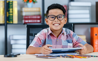 Smart Asian boy wearing glasses and pink checked shirt sitting on desk of magnifying glass, smartphone, and colorful pencils in reading room and concentrate on tablet study to complete homework