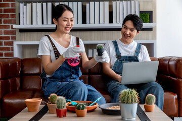 Young asian couple wearing jean overall and white t-shirt sitting at home while taking care of small cactus and using laptop. Concept home gardening hobby