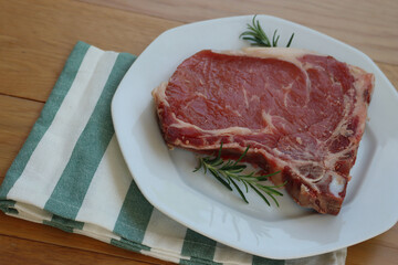 Raw beef meat steak on a white plate on wooden table with fresh rosemary herbs ready to cook