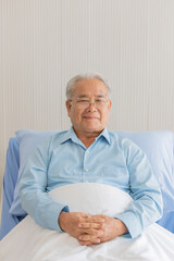 Close up portrait shot of an old fat asian patient with gray hair wearing eyeglasses and light blue long sleeve shirt sitting on hospital bed cover his lower body with white clean blanket