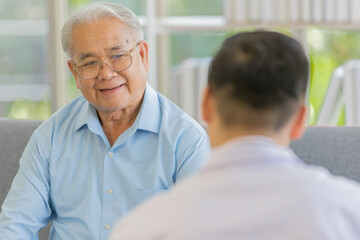 An old fat male patient with gray white hair wearing light blue shirt and brown pants sitting on sofa and discussing about health with a young doctor with black hair wearing white lab coat