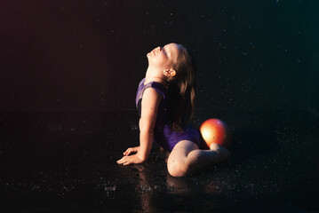 girl gymnast in purple swimsuit does exercises with a ball on a black background in the water, advertising sports goods - Powered by Adobe