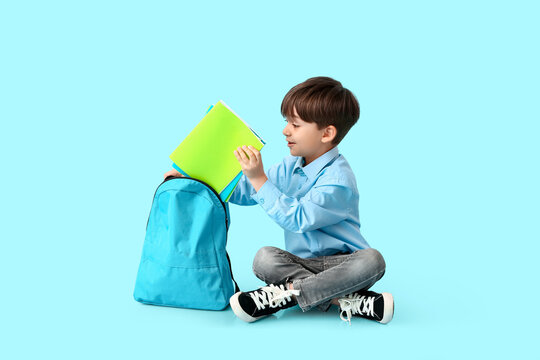 Little schoolboy putting notebooks in backpack on color background