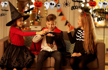 halloween, holiday and childhood concept - happy smiling boy and girls in costumes sharing candies at home at night decorated with garland and lights
