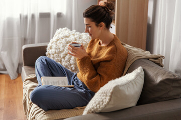 people and leisure concept - young woman drinking coffee and reading book sitting on sofa at home