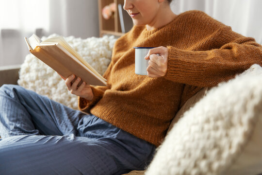 people and leisure concept - young woman drinking coffee and reading book sitting on sofa at home
