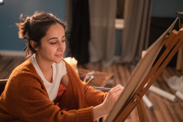 Portrait of an attractive young female drawing pictures at night in her home's cozy living room. The woman smiles resting her hand with a pencil on a drawing block leaning against a canvas © ABCreative