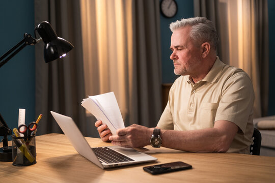 An Elderly Man With Impaired Vision Tries To Read Documents, Reads Invoices, Counts Document Pages. Concerned Senior Checks Bank Statements. Focused Elderly Man Sits At Laptop And Pays Bills Online.