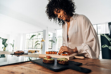 Fototapeta na wymiar Young african american woman cutting tomatoes preparing breakfast avocado toast in bright loft kitchen