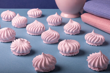 Beautifully arranged rows of delicate pink meringue cookies on a light blue background next to a harmonious pink and blue cloth napkins and a pink large cup. Light dessert.