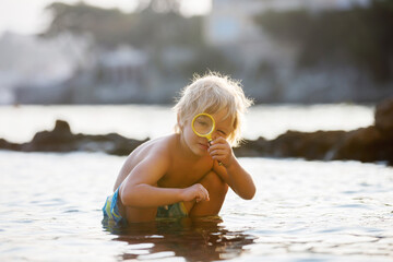 Cute child, boy, using magnifying glass to examine seashells on the beach