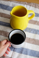 Black coffee in white enamel mug in hand and yellow ceramic jug on a linen striped tablecloth