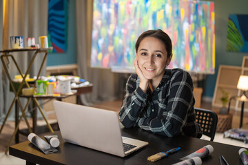 Portrait of smiling brunette wearing a checkered shirt sitting at a desk in front of a laptop in...