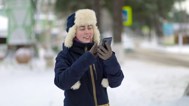 Portrait of a young smiling woman in a hat with earflaps and a jacket, who takes a selfie on her smartphone. Snow-covered street in the background.