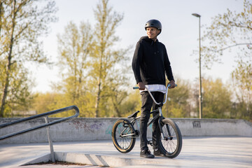 Obraz premium A teenager stands in a helmet with his bike dressed in casual street style on a platform at a skatepark. The boy is looking at a group of girls, smiling and waiting for them to come up