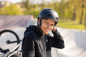 A boy sits on a ramp at a skatepark with his bike lying wheel up behind him. The guy has finished riding and removes his black helmet from his head, unzips it behind his head