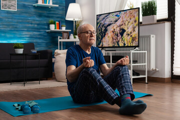 Senior man sitting comfortable in lotus position on yoga mat with closed eyes training body...