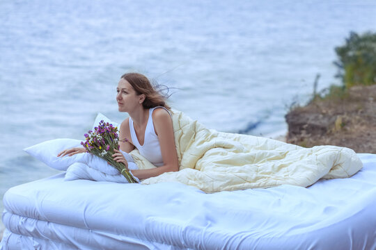 The bed is on the ocean. On the bed is a beautiful woman, in her hands a bouquet of flowers. The woman on the bed looks at the ocean.