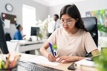 Busy woman of Asian Korean beauty dressed in loose T-shirt and glasses sits at desk in front of computer working on new project, she holds piece of paper and pen in front of her drawing patterns