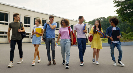 Obraz premium Group of diverse young people in casual wear with backpacks on shoulders and books in hands walking in slow motion against college building and talking. Tracking shot students after classes