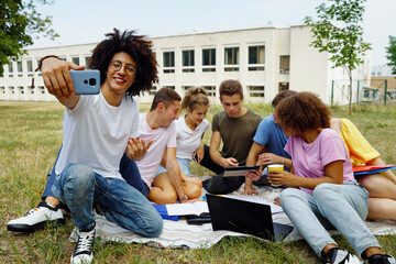 Black man recording video while his multiethnic friends sitting together on blanket and using tablet for studying, college building on background. Blogger streaming online from campus