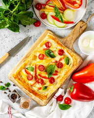 Puff pastry pie with mixed vegetables and mozzarella cheese on a serving board on a culinary light table top view. Delicious hearty pastries with tomatoes, zucchini, sweet pepper, spices and basil