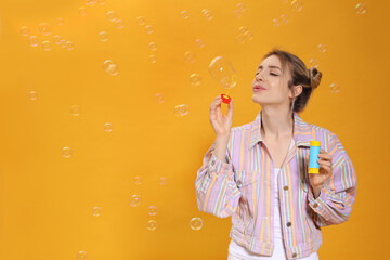 Young woman blowing soap bubbles on yellow background, space for text