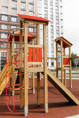 An outdoor colorful playground next to the house. A summer day. Children's playground with rubber floor covering. A wooden house with a slide and ropes.
