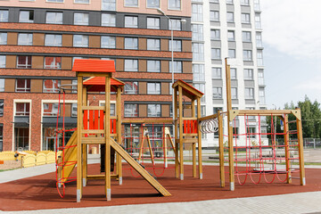 Fototapeta na wymiar An outdoor colorful playground next to the house. A summer day. Children's playground with rubber floor covering. A wooden house with a slide and ropes.