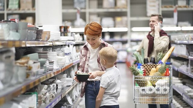 Family with child boy choosing goods in supermarket, store, caucasian redhead woman with son looking for the best kitchen utensils, discussing. Husband talk on phone in the backgorund. Shopping