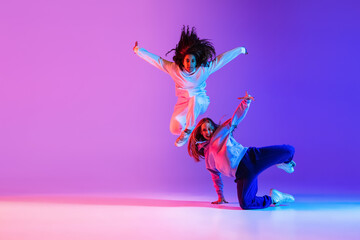 Two active young girls dancing hip-hop on gradient pink purple neon studio background