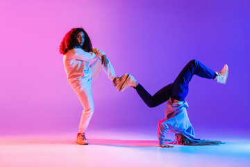 Two beautiful young girls dancing hip-hop dynamically on colorful gradient background in neon.