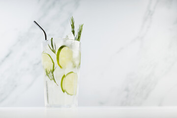Fresh citrus infused homemade lemonade with ice cubes, lime slices, green rosemary twig, silver straw in soft light white kitchen interior in sunny day.