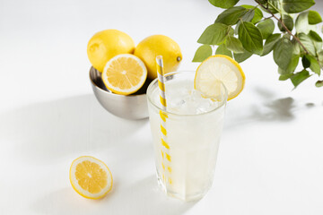 Detox fresh lemon juice of ripe lemons with ice in glass with ingredients in silver bowl, green branch in summer sunlight on white table.