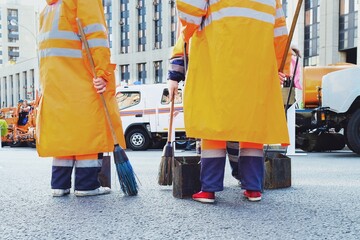 The back view close up of municipal workers in orange uniform sweep city streets with manual cleaning equipment. Urban sweepers team with broomsticks and scoops for garbage on blurred city background.
