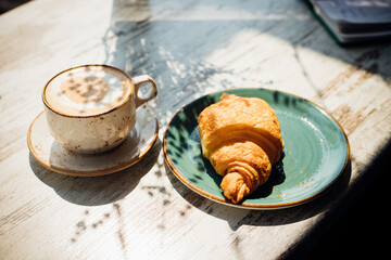 Cappuccino and croissant are on the table in the cafe. Morning sunlight falls on the table, beautiful shadows appear.