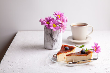 Slices of Homemade Basque burnt cheesecake on plate with blueberries and mint leaves, cup of black tea on light background with flowers in vase