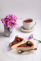 Slices of Homemade Basque burnt cheesecake on plate with blueberries and mint leaves, cup of black tea on light background with flowers in vase