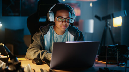 Young Teenage Multiethnic Girl Using Laptop Computer and Wearing Headphones in a Dark Cozy Room at Home. She's Browsing Educational Research Online. Studying Science School Homework Concept.