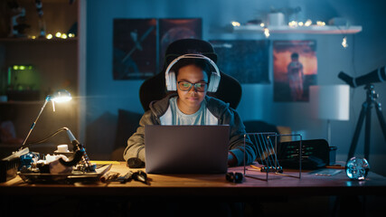 Young Teenage Multiethnic Girl Using Laptop Computer and Putting On Headphones in a Dark Cozy Room at Home. She's Browsing Educational Research Online. Studying Science School Homework Concept.