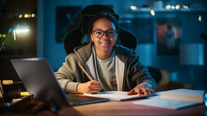 Fototapeta na wymiar Young Teenage Multiethnic Black Girl Writing Down Homework in a Notebook with a Pencil, Using Laptop Computer in a Dark Cozy Room at Home. She's Happy, Looks at Camera and Smiles.