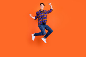 Fototapeta na wymiar Photo of sweet lucky teenager man dressed checkered clothes jumping high rising fists smiling isolated orange color background