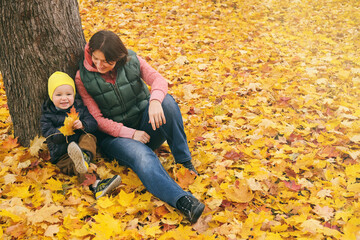 Mother and her little child boy sitting near huge maple tree in autumn park and looking on falling leaves. Fall nature banner with copy space.