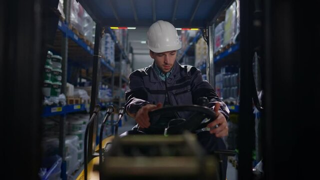 Portrait of concentrated Caucasian young man in hard hat turning on forklift sitting in vehicle indoors. Front view of focused Caucasian warehouse loader using transportation in storehouse