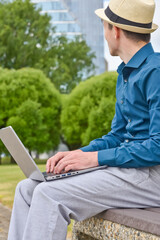 A man with a laptop in the summer works remotely outdoors and looks towards the office building.