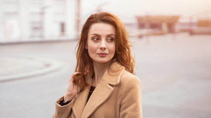 Beautiful woman standing outdoors posing for camera in an autumn beige coat, beautiful hair with urban city background. Vertical portrait. Warm tinted photo. 