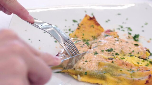 Juicy casserole with spinach and poured salmon cream on white plate in luxury restaurant, person hands using fork and table knife for cutting baked seafood pudding on pieces. Baked marine casserole