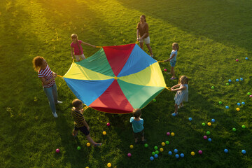 Group of children and teachers playing with rainbow playground parachute on green grass, above...